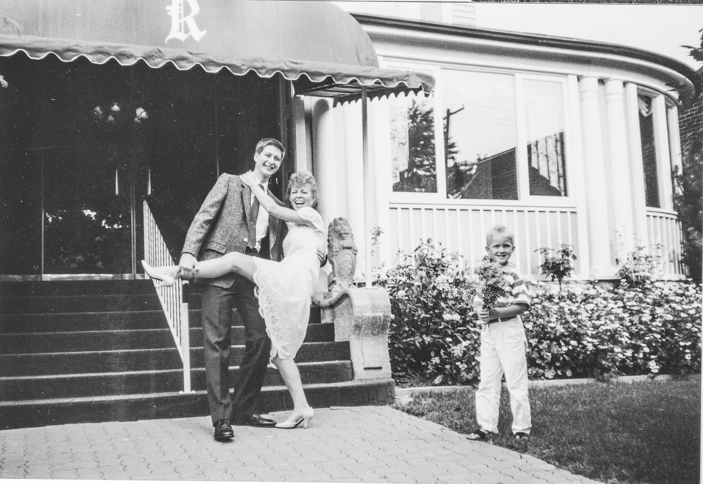 black and white image of Lina laughing while a man holds her leg up and her son grins with flowers at the fake wedding she put on to prank a friend